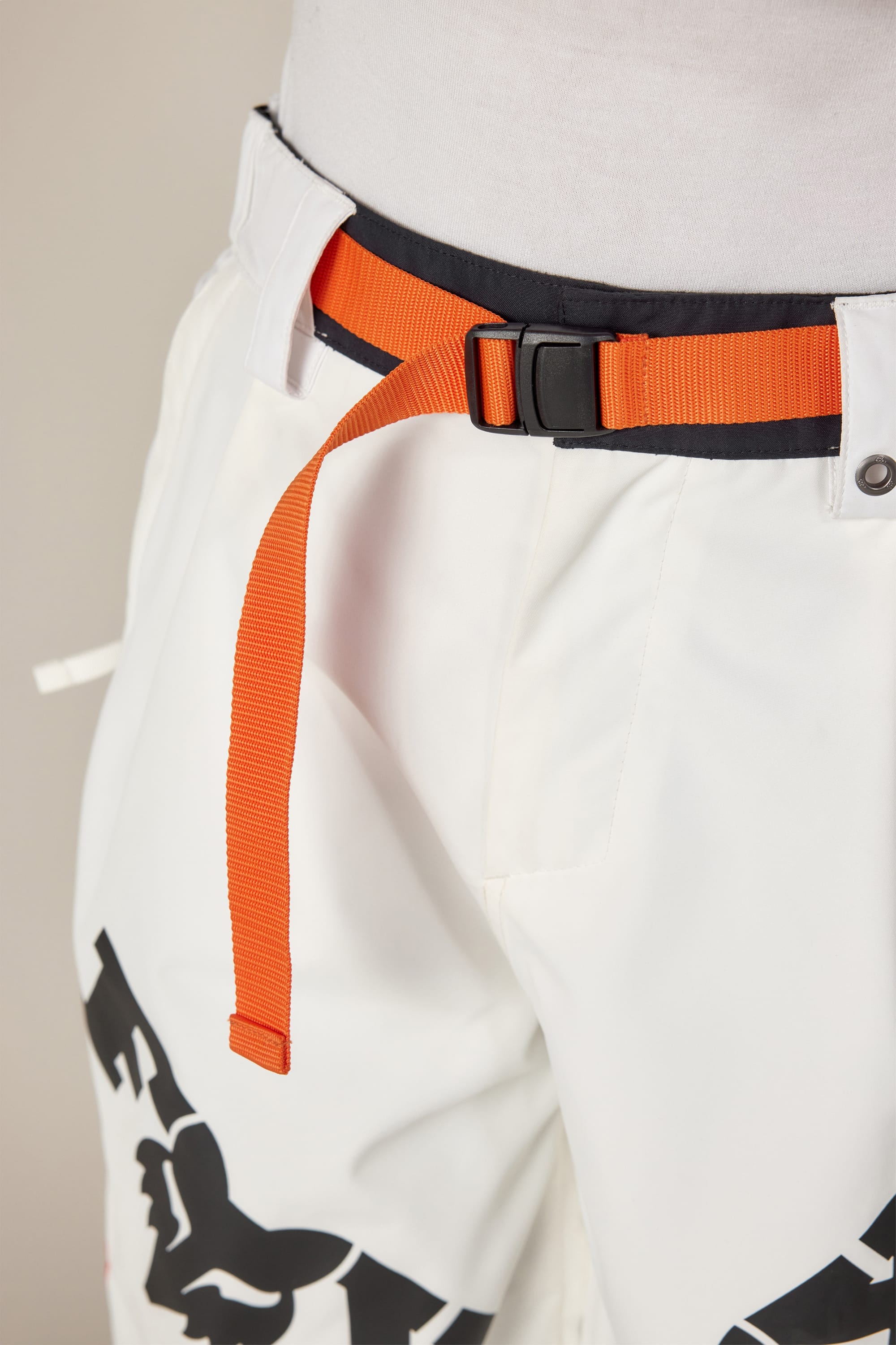A close-up of a person wearing 686 Women's Fox Racing Shell Pant in white with black graphic designs and an orange webbing belt with a black buckle, fastened loosely around the waist.