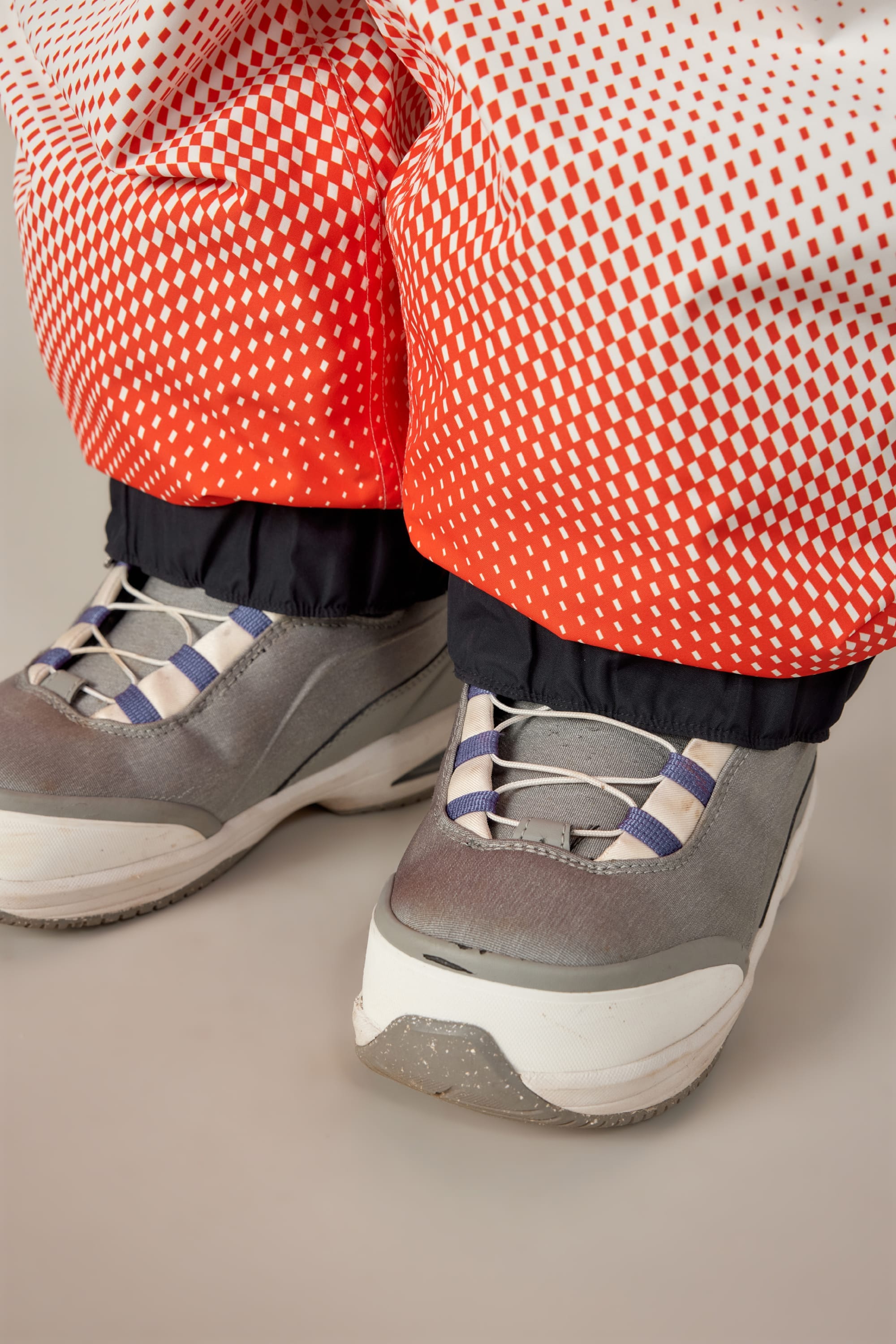 Close-up of a person wearing gray sneakers with white soles and purple-striped laces, paired with 686 Women's Fox Racing Shell Pants in red and white, tucked into the tops of the shoes.