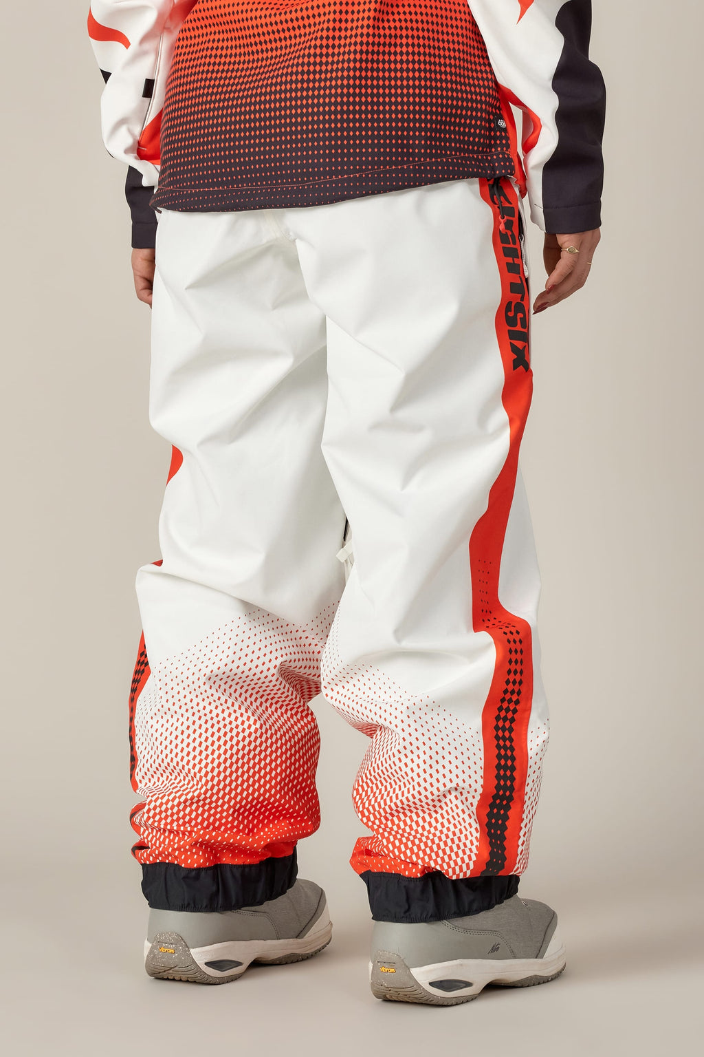 A person is seen from behind wearing 686 Women's Fox Racing Shell Pant by 686, featuring white fabric with red gradient dots and "ANTISOCIAL" on the side, paired with light gray sneakers against a plain background.