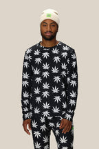 A man stands facing forward in a 686 Men's HUF Plantlife Base Layer LS Top featuring a white cannabis leaf pattern, paired with matching pants. He completes the look with a white beanie with a green patch against a plain beige background.