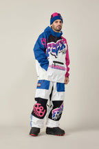 A man stands against a plain background wearing the 686 Men's Fox Racing Waterproof Hoody—blue, pink, and white with bold graphics—paired with matching baggy pants, a blue-pink beanie, and black snow boots.