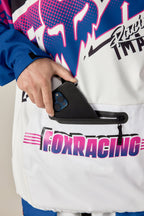A person wearing a 686 Men's Fox Racing Waterproof Hoody with bold pink and blue graphics puts a black smartphone into the jacket’s front zippered white pocket.