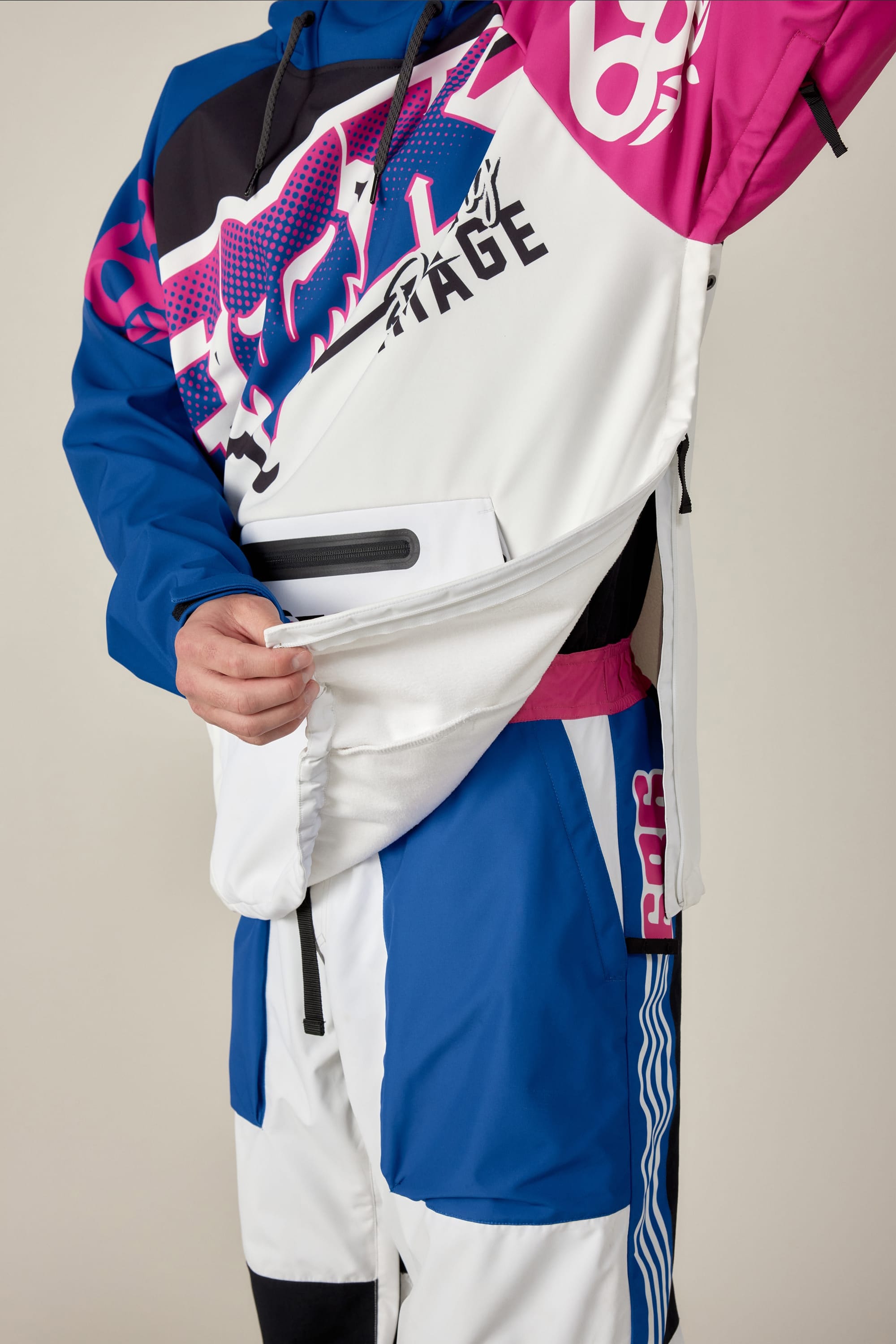 A person in the 686 Men's Fox Racing Waterproof Hoody shows the large side zipper being unzipped to reveal an inner pocket. The hoody features blue, white, pink, and black patterns; only the torso and part of the legs are visible.