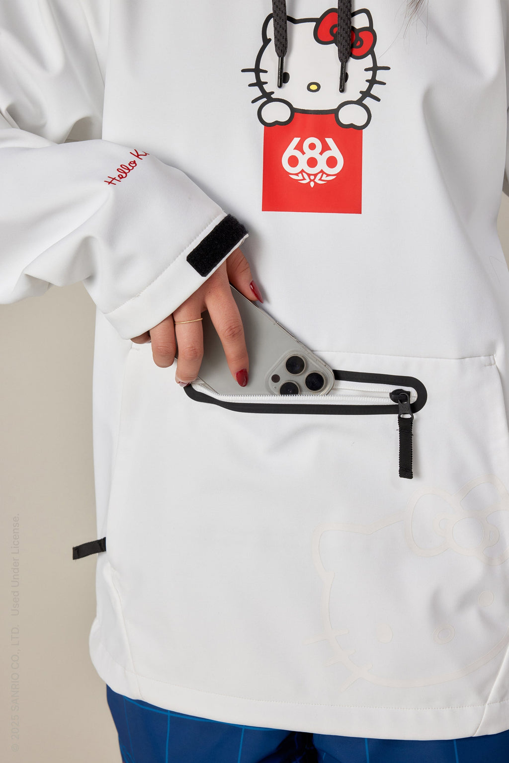 A person wearing a 686 Women's Hello Kitty® Waterproof Hoody in white, featuring a red Hello Kitty logo, places a silver smartphone into the front zippered pocket. Only their torso and hand are shown. Brand: 686.