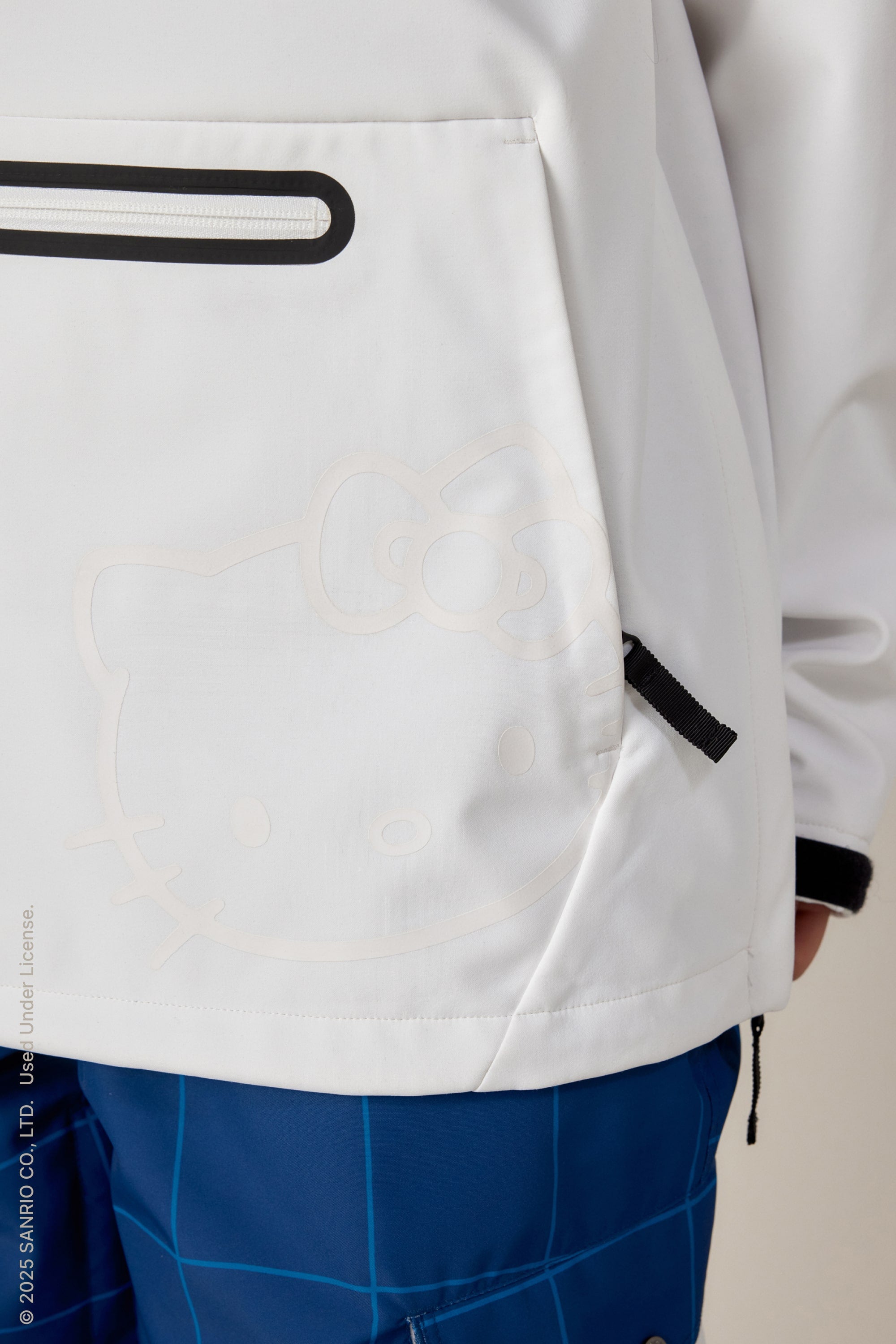 A close-up of a person wearing the 686 Women's Hello Kitty® Waterproof Hoody, featuring a faint Hello Kitty face design on the front pocket, paired with blue checkered pants. The person's hand is partially visible on the right.