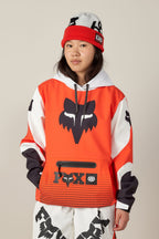 A young woman wears the 686 Women's Fox Racing 97 Waterproof Hoody and matching pants, plus a red and gray beanie. She stands against a plain background, looking at the camera with a neutral expression.