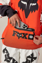 A person in a 686 Women's Fox Racing 97 Waterproof Hoody places a gray smartphone into an orange FOX waist bag, matching their outfit. Their nails are painted red.