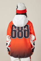 A person faces away, wearing the 686 Women's Fox Racing 97 Waterproof Hoody in orange with bold black "686" numbers on the back, paired with a white and red beanie. The background is plain and neutral.