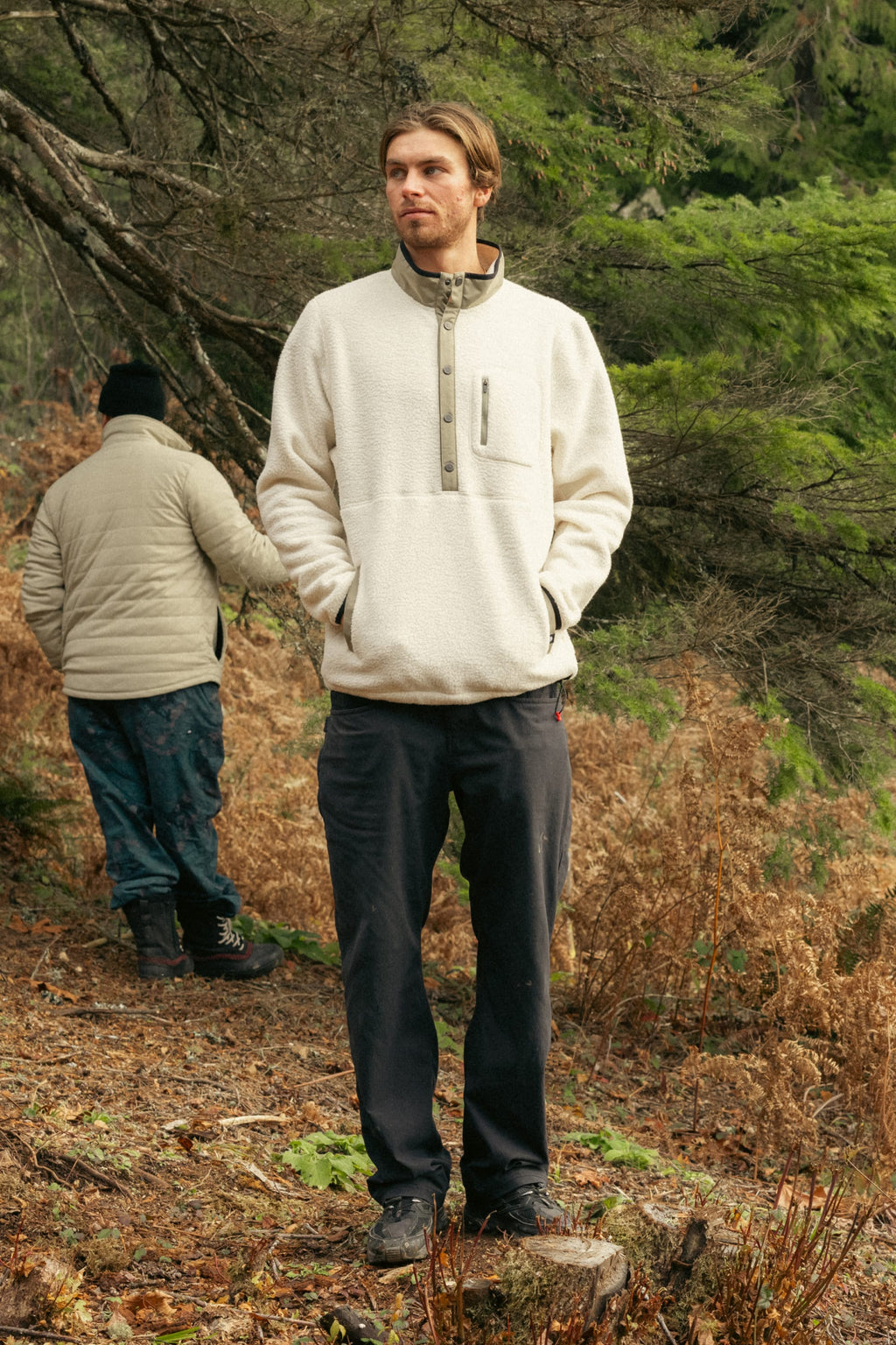 A man in a light beige 686 Mens Tioga Fleece Pullover and dark pants stands outdoors in a forested area with his hands in his pockets. Another person is seen turned away among trees and ferns in the background.