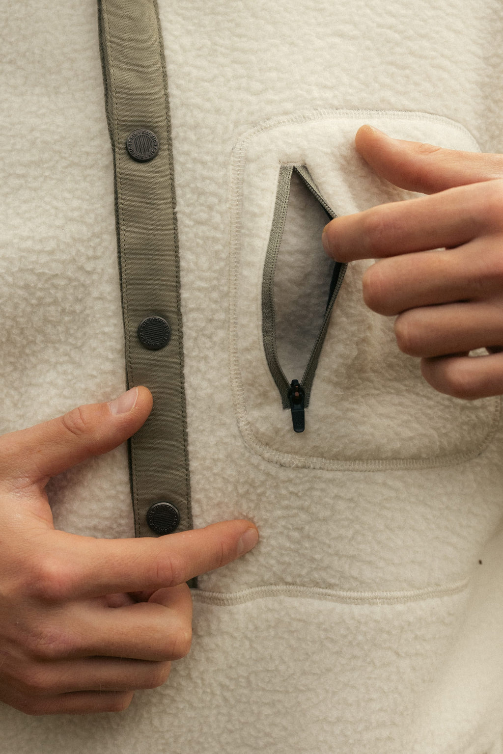 A man opens a small zippered chest pocket on the cream-colored 686 Mens Tioga Fleece Pullover, featuring dark buttons and trim.