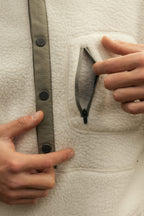 A man opens a small zippered chest pocket on the cream-colored 686 Mens Tioga Fleece Pullover, featuring dark buttons and trim.