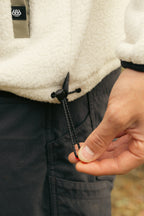 A person adjusts the hem drawstring of a white 686 Mens Tioga Fleece Pullover, pulling the black cord with their right hand. They are also wearing dark pants.
