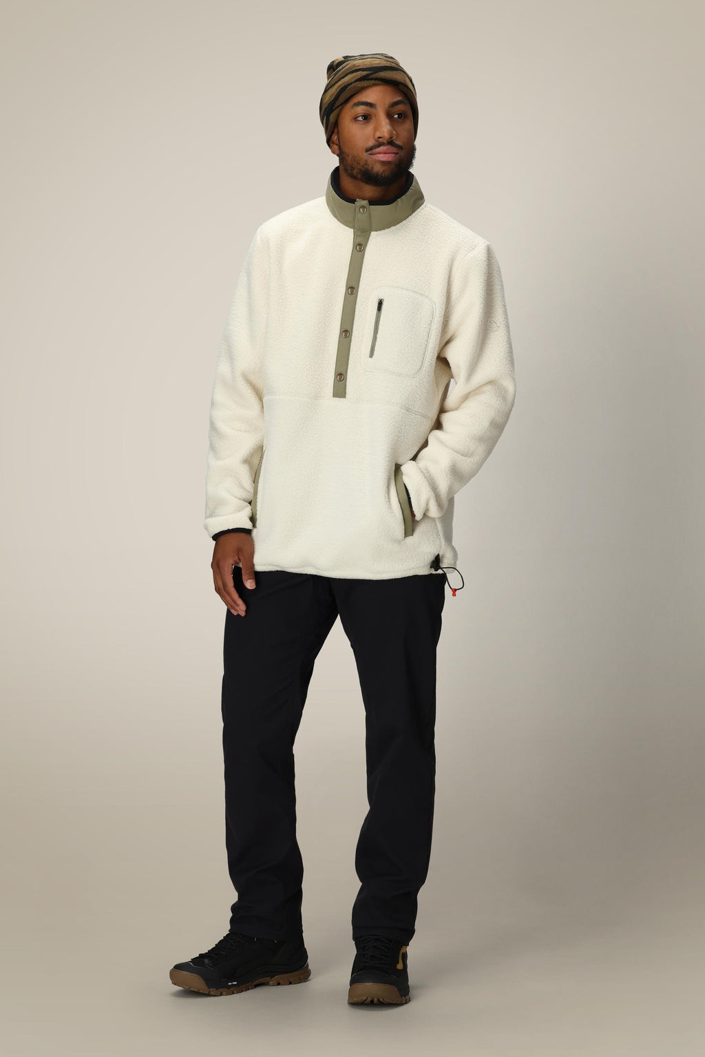 A man in a 686 Mens Tioga Fleece Pullover, black pants, brown hiking shoes, and a striped beanie stands against a neutral background with one hand in his pocket.