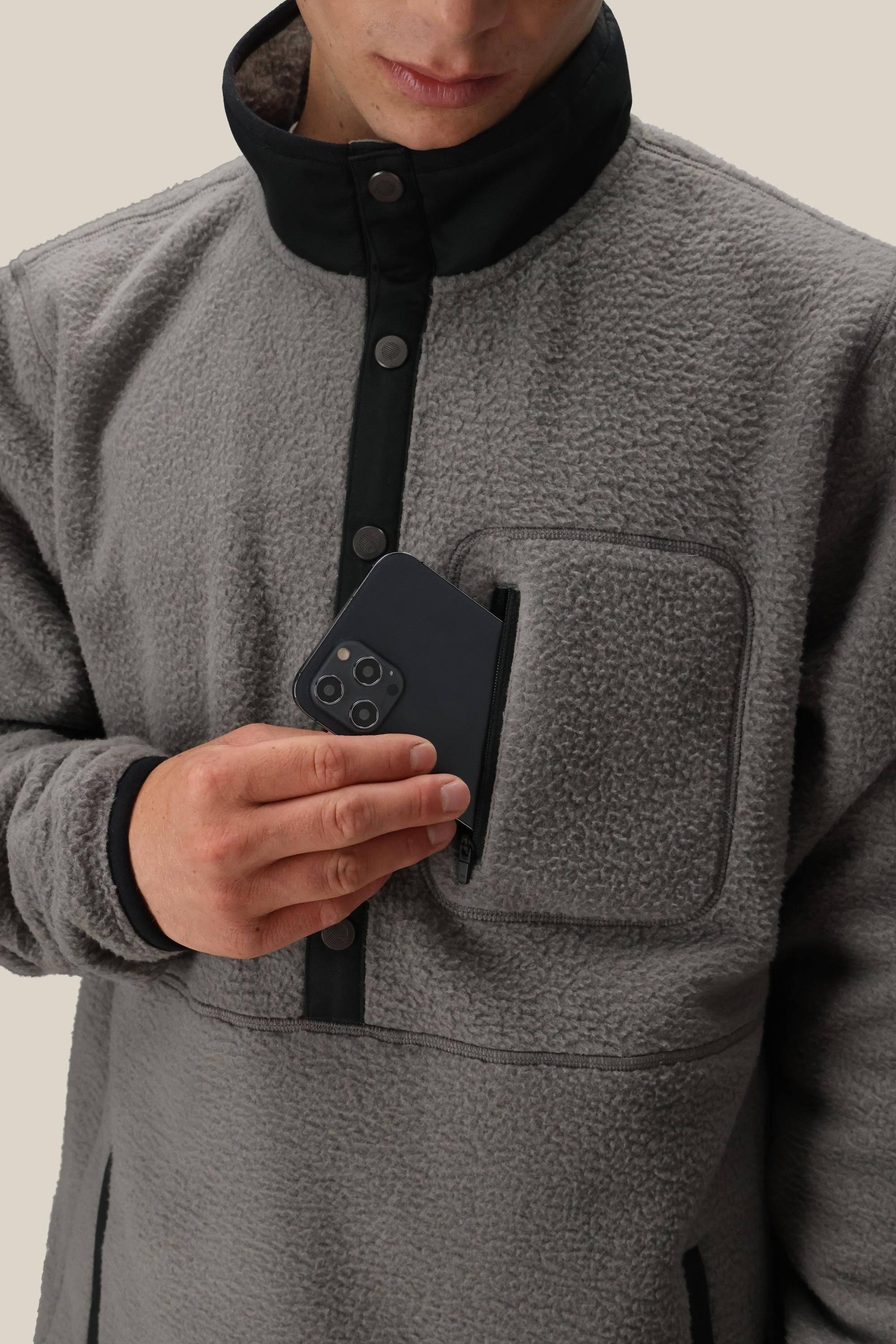 A man in a 686 Mens Tioga Fleece Pullover (grey, polyester, high collar) places a black smartphone into the chest zipper pocket. The background is plain and light-colored.