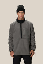 A man wears a 686 Mens Tioga Fleece Pullover by 686 with black buttons and a zippered chest pocket, paired with a black beanie, standing against a plain light beige background.