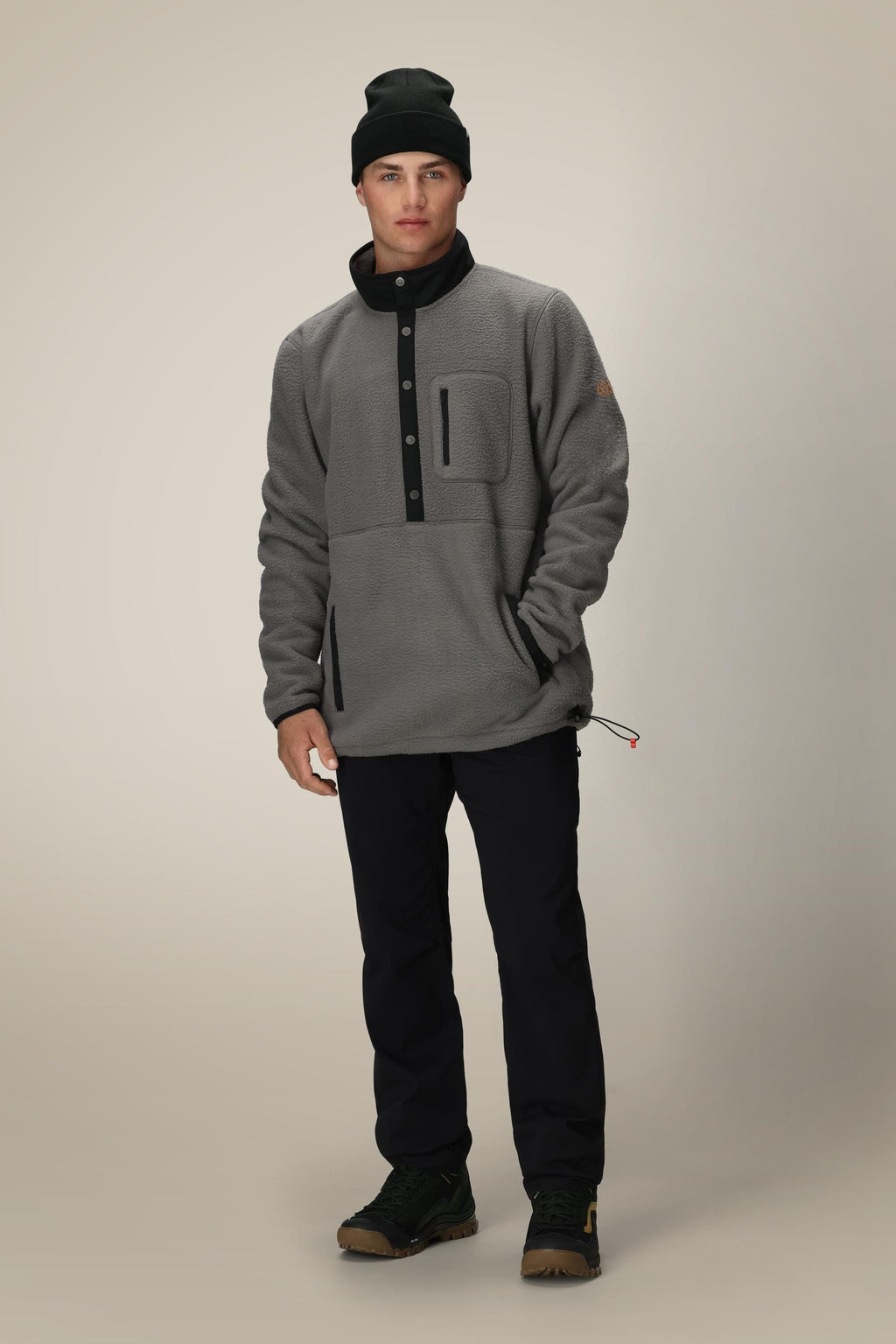 A man faces forward against a neutral background, wearing a black beanie, black pants, brown hiking boots, and the 686 Mens Tioga Fleece Pullover by 686—a grey polyester sweatshirt pullover with buttons.