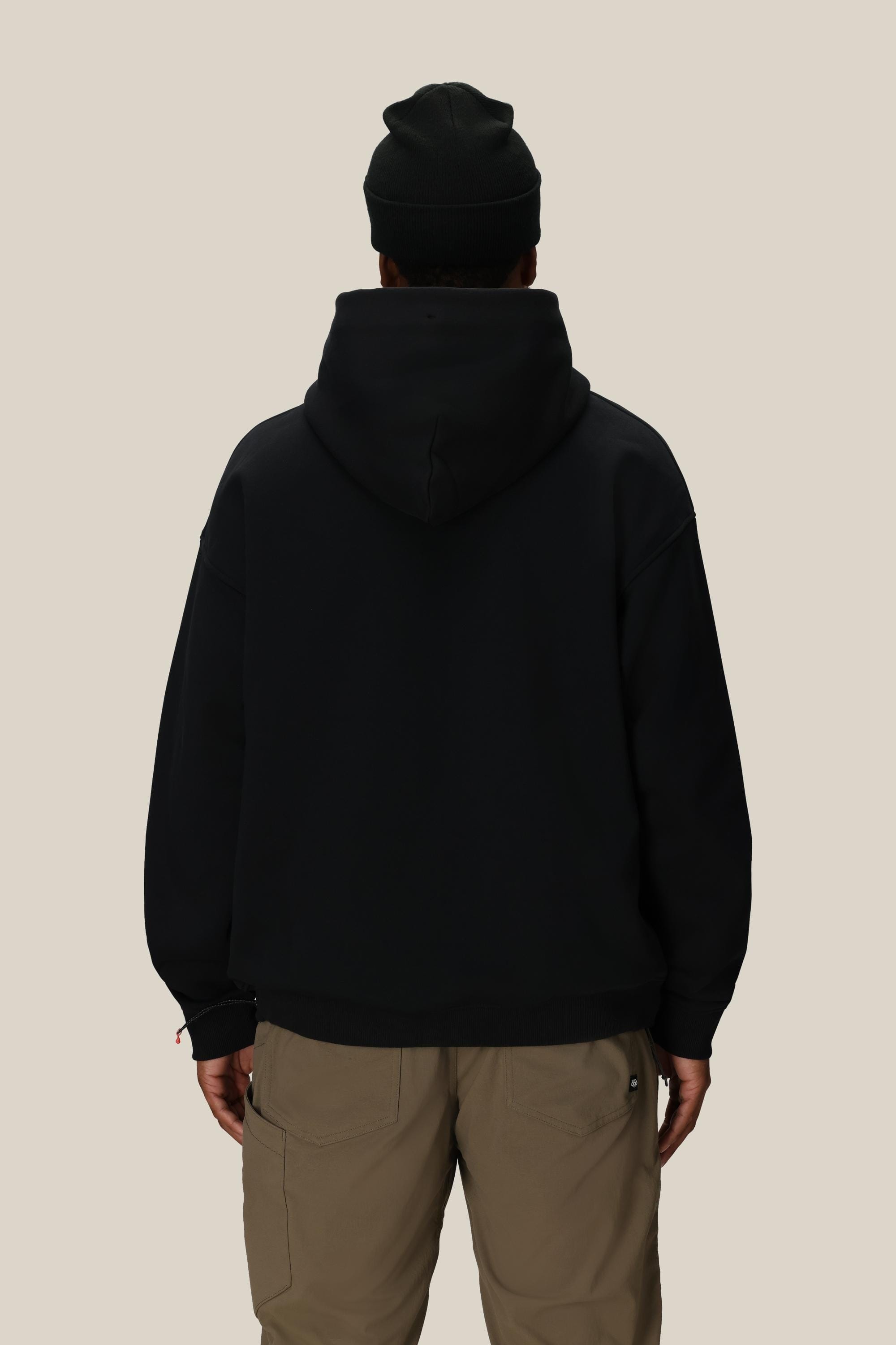 A person stands facing away from the camera, hands by their sides, wearing a black 686 Knockout Logo Premium Heavyweight Pullover Hoody, a black beanie, and light brown pants against a plain beige background.