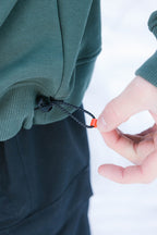 A close-up of someone adjusting the black drawstring with an orange toggle on the hem of a green 686 Knockout Logo Premium Heavyweight Pullover Hoody, paired with black pants against a snowy white background.