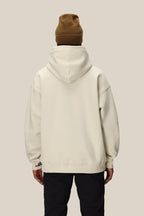 A person wearing a brown beanie, black pants, and the 686 Knockout Logo Premium Heavyweight Pullover Hoody by 686 stands with their back to the camera against a plain beige background.