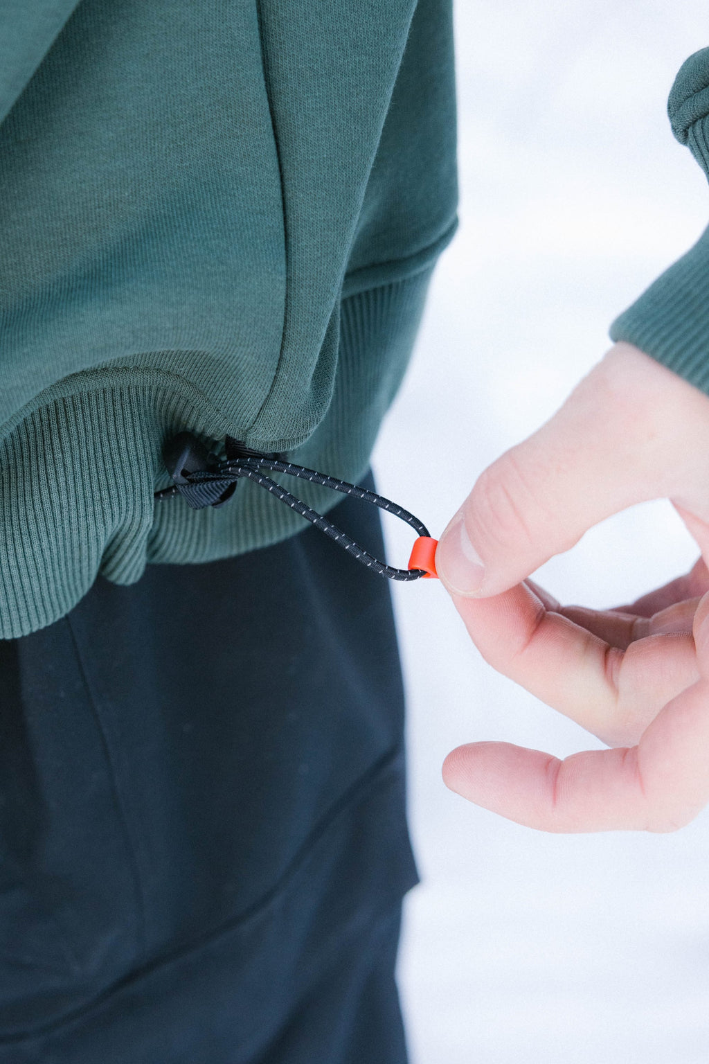 A person tightens the drawstring hem of a green 686 Knockout Logo Premium Heavyweight Pullover Hoody with their right hand. They’re wearing black pants, and the blurred white background suggests snow.