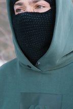 A person wearing a 686 Knockout Logo Premium Heavyweight Pullover Hoody in green and a black knit face mask that covers their nose, mouth, and chin, leaving only their eyes and part of their face visible.