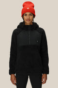 A person faces forward against a plain beige background, wearing the 686 Womens Hemlock Fleece Hoody in black and a bright red knit beanie.