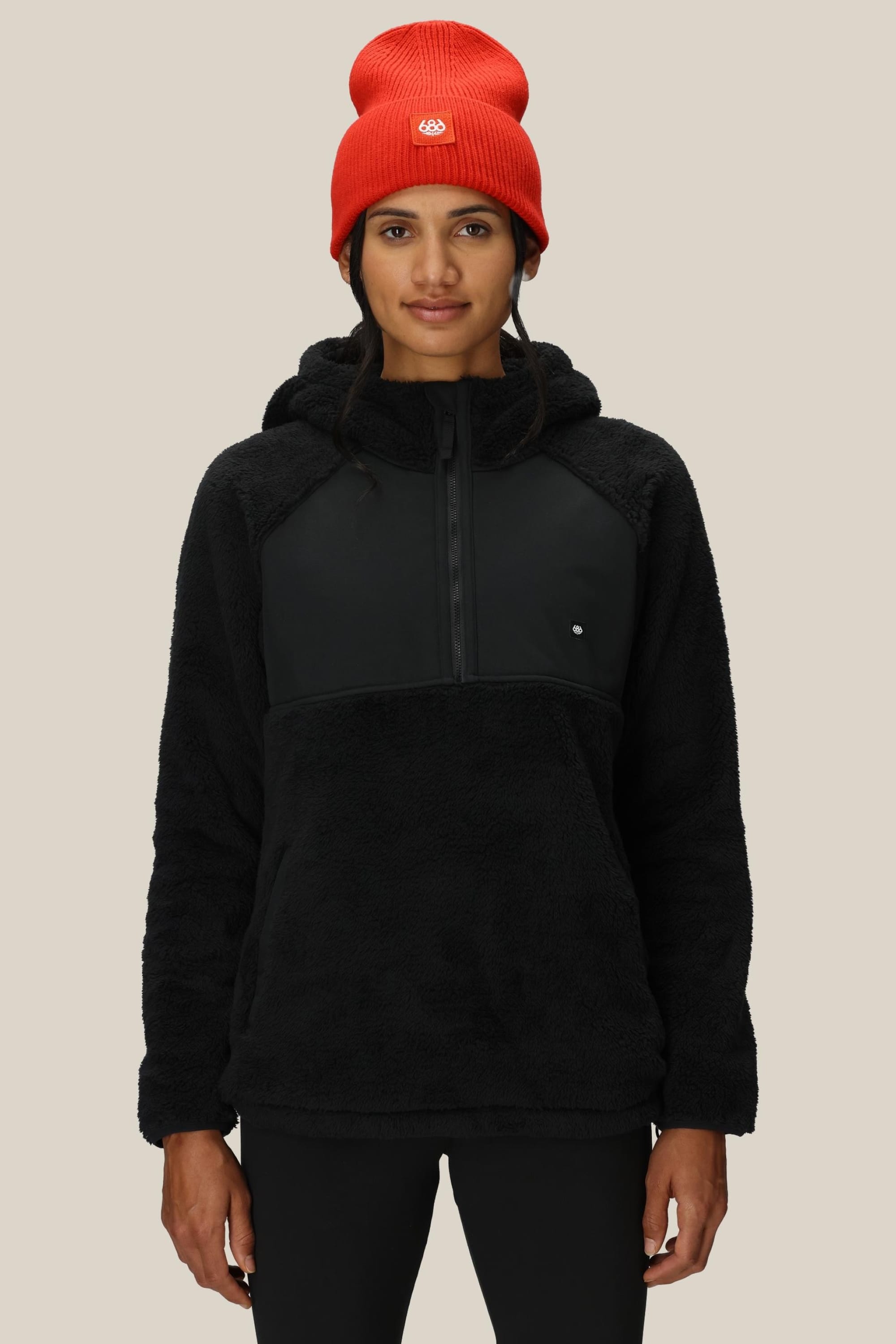 A person faces forward against a plain beige background, wearing the 686 Womens Hemlock Fleece Hoody in black and a bright red knit beanie.