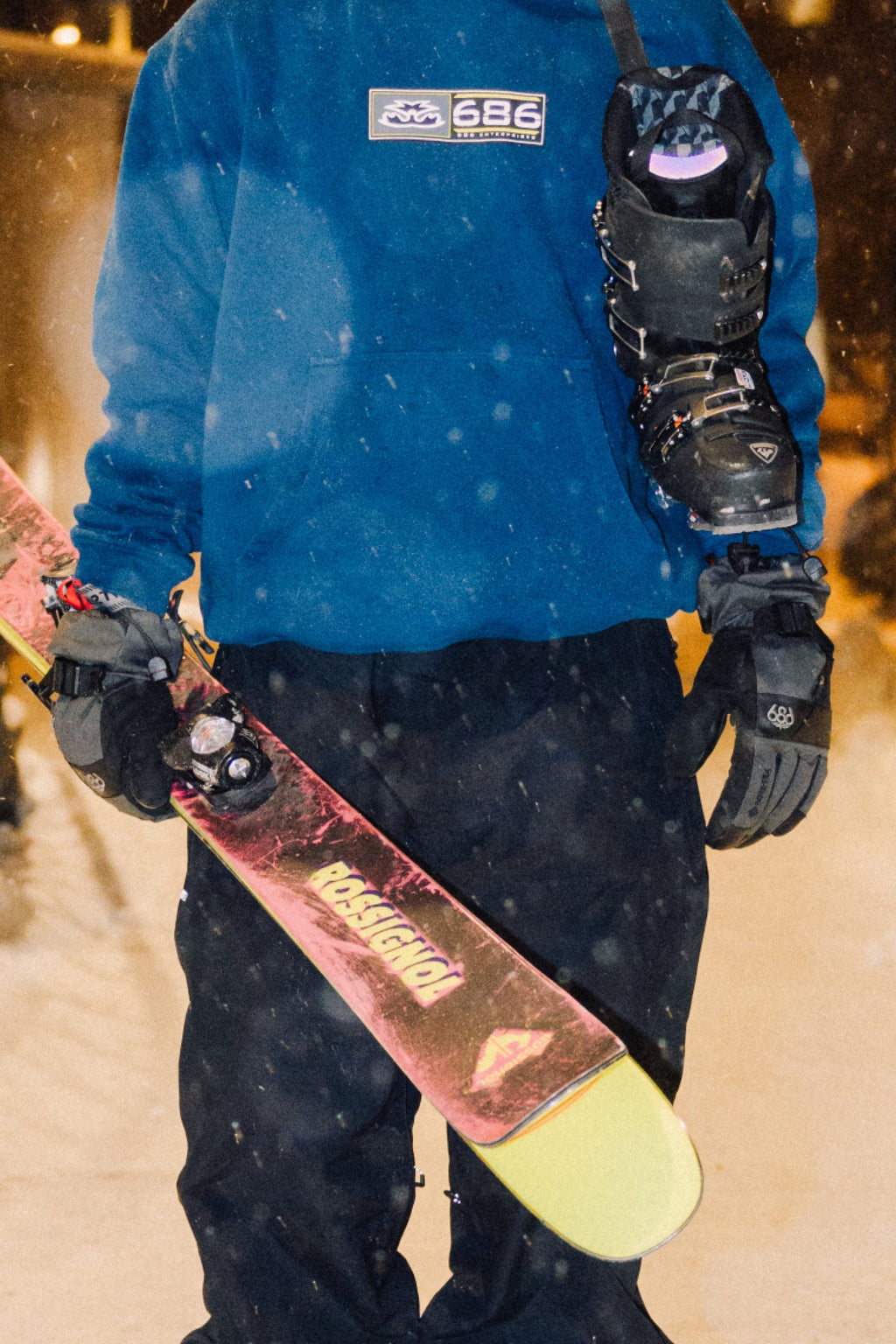 Wearing a blue sweatshirt and 686 Men's GORE-TEX SMARTY® 3-In-1 Gauntlet Gloves, a person holds colorful skis with a snowboard boot over their shoulder, standing outside on a snowy night.