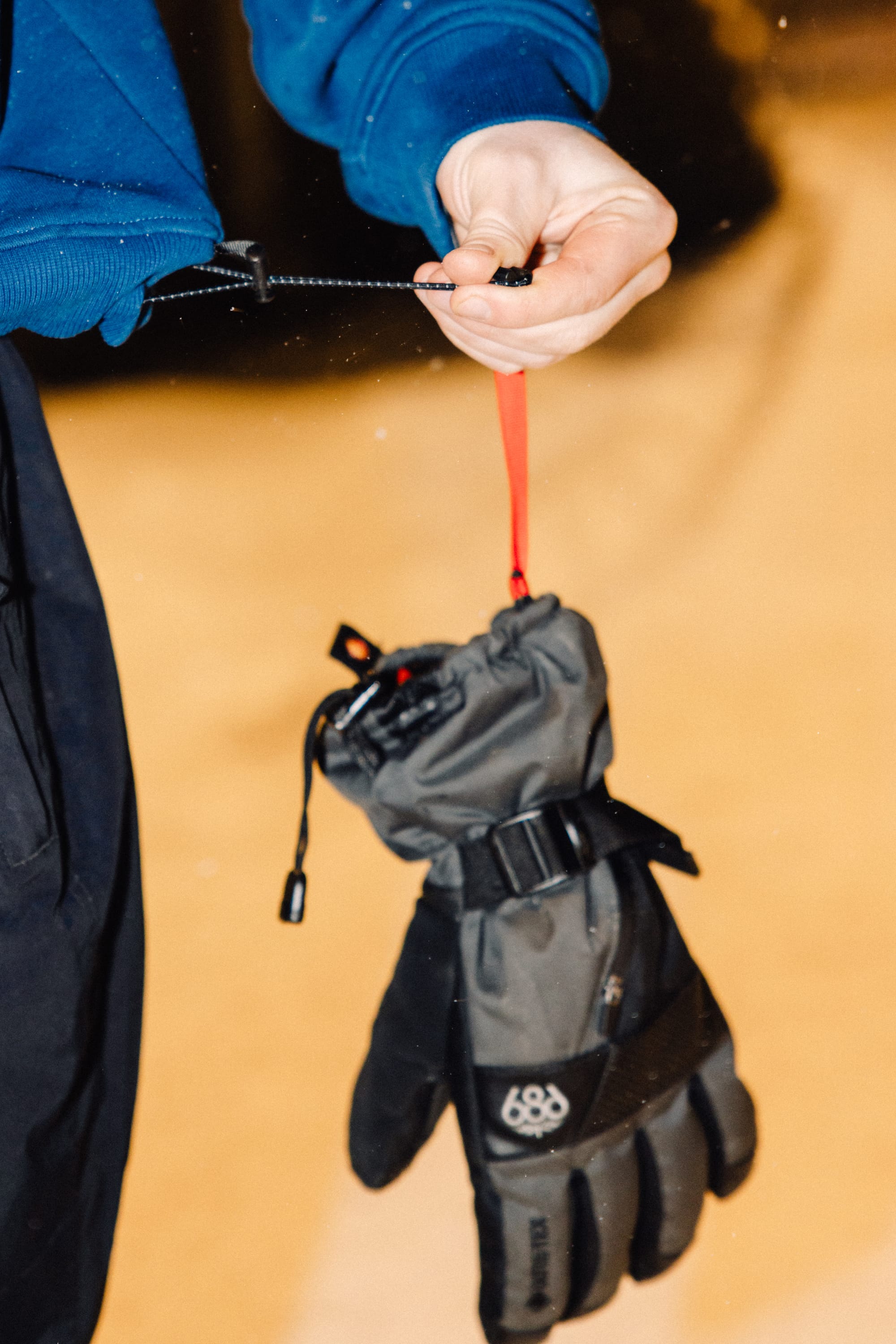 A person in a blue jacket holds a 686 Men's GORE-TEX SMARTY® 3-In-1 Gauntlet Glove by its red strap. The glove, from 686, hangs down against an out-of-focus light background that may be snow.