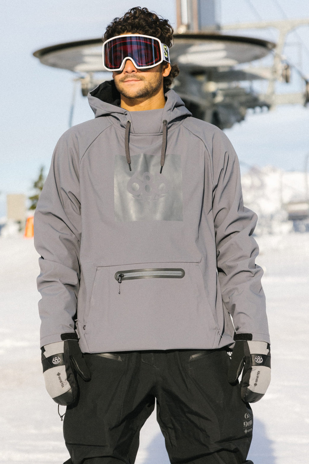 A man stands on snow with a ski lift in the background, wearing a gray hooded ski jacket, black pants, goggles, and 686 Men's GORE-TEX SMARTY® 3-In-1 Gauntlet Mitts by 686.