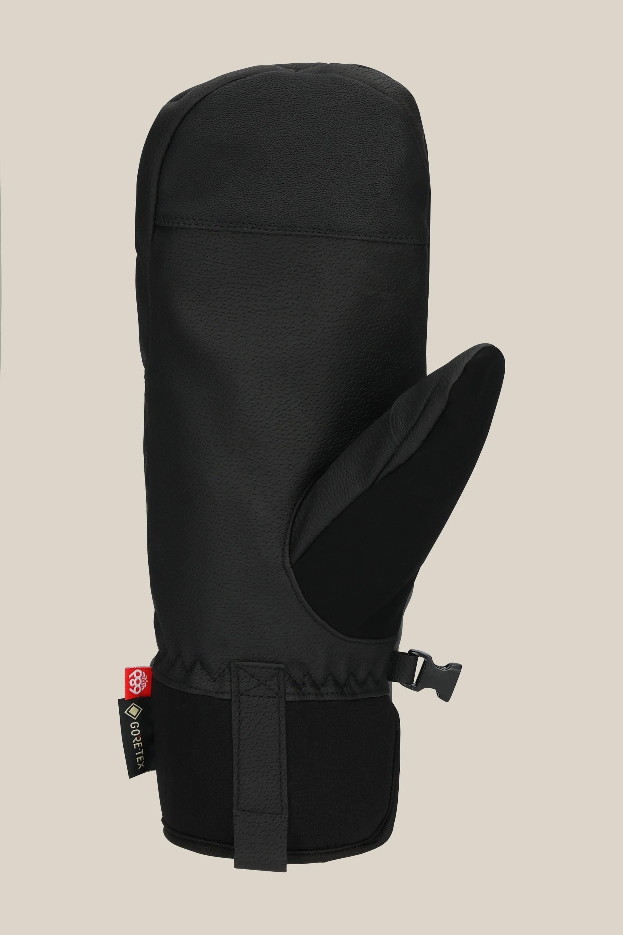 The 686 Men's GORE-TEX Linear™ Under Cuff Mitt in black features a textured surface, adjustable wrist strap, and a red-green tag, shown against a beige background. This durable PU mitt from 686 is stylish and ideal for winter wear.