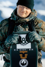 A person in camo winter gear and 686 Men's GORE-TEX Linear™ Under Cuff Mitts holds a snowboard with 686 logos, smiling outside in the snow and wearing a black beanie.