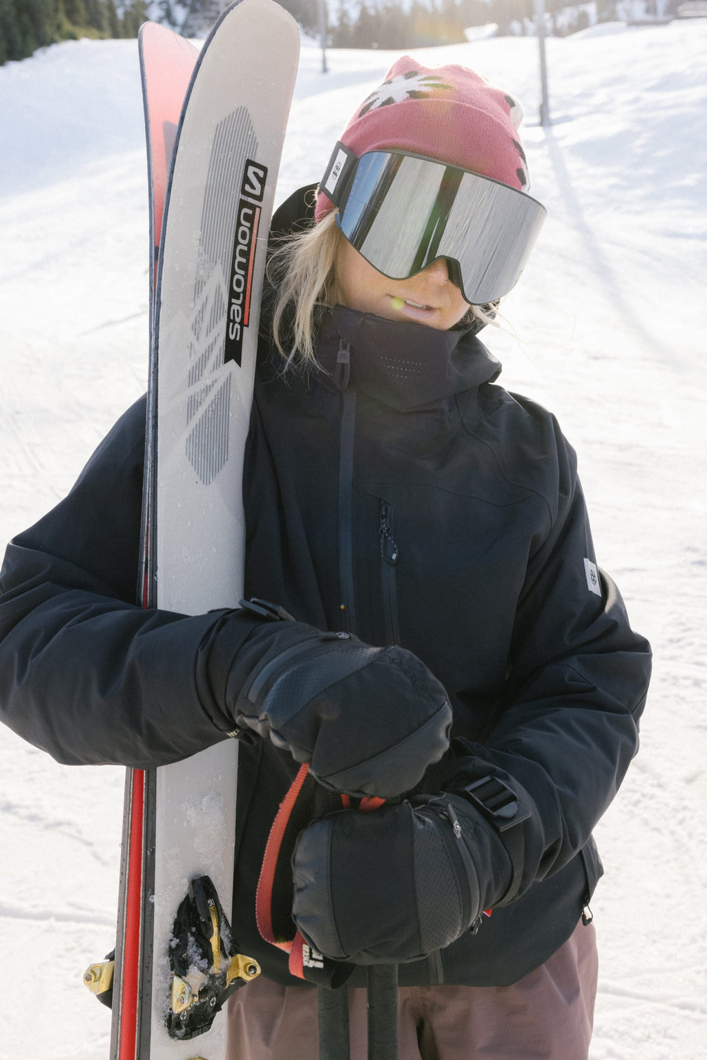 A person in a pink beanie, ski goggles, and black jacket stands on a snowy slope holding Salomon skis upright. They're wearing 686 Women's GORE-TEX SMARTY® 3-In-1 Gauntlet Mitts by 686 as the sun shines brightly behind them.
