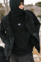 Wearing the 686 Mens Clava Fleece Hoody, a person stands outdoors on a snowy day. Dressed in black winter gear, they look to the side with blurred trees and snow in the background.