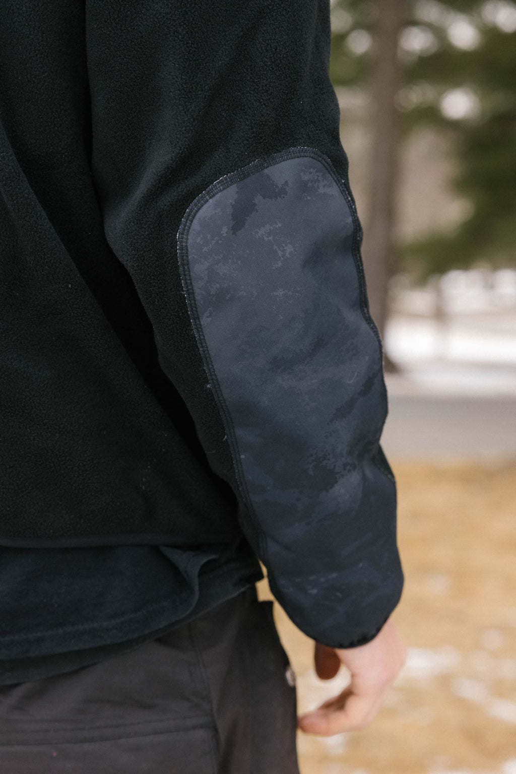 A close-up of a persons arm wearing the 686 Mens Clava Fleece Hoody, featuring a black polyester design with a dark reinforced elbow patch, outdoors on a wintry day with blurred trees and snow in the background.