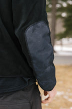 A close-up of a persons arm wearing the 686 Mens Clava Fleece Hoody, featuring a black polyester design with a dark reinforced elbow patch, outdoors on a wintry day with blurred trees and snow in the background.