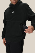 A person wearing the 686 Mens Clava Fleece Hoody in black, featuring a small white 686 logo, slips a black smartphone into its front kangaroo pocket against a plain beige background.