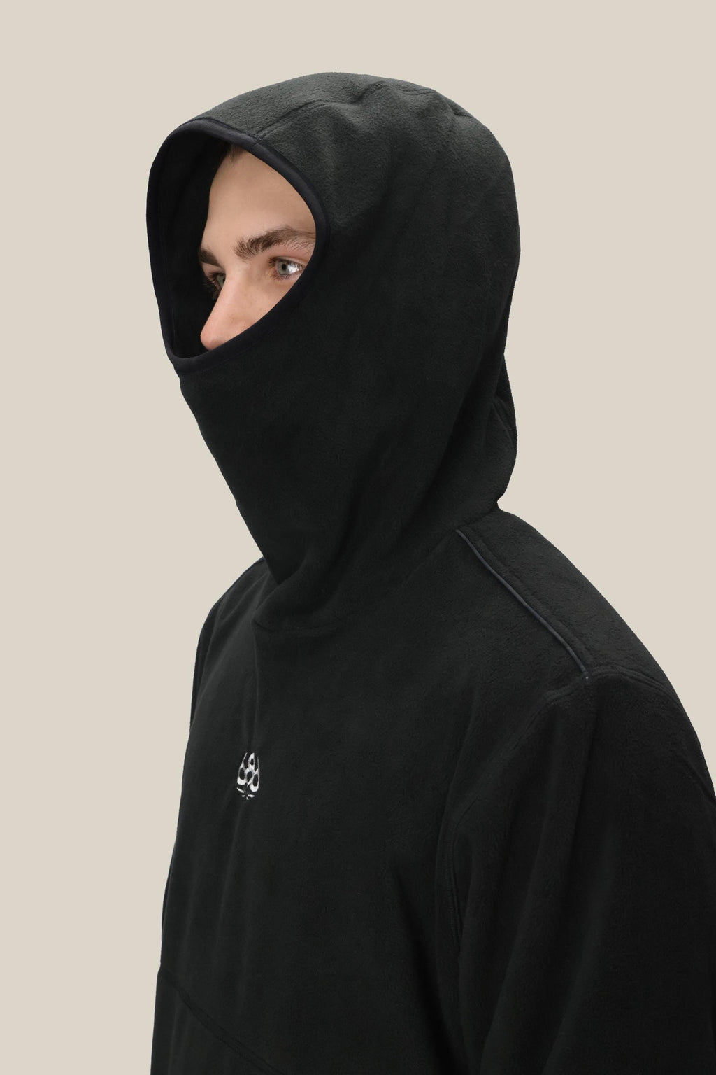 A person wearing the 686 Mens Clava Fleece Hoody by 686, its black hood obscuring all but their eyes and upper nose, stands against a plain beige background.