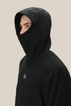 A person wearing the 686 Mens Clava Fleece Hoody by 686, its black hood obscuring all but their eyes and upper nose, stands against a plain beige background.