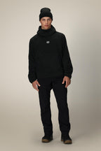 A young man stands facing forward, wearing the 686 Mens Clava Fleece Hoody in black with a small white logo, paired with black pants and brown shoes, set against a plain light beige background.