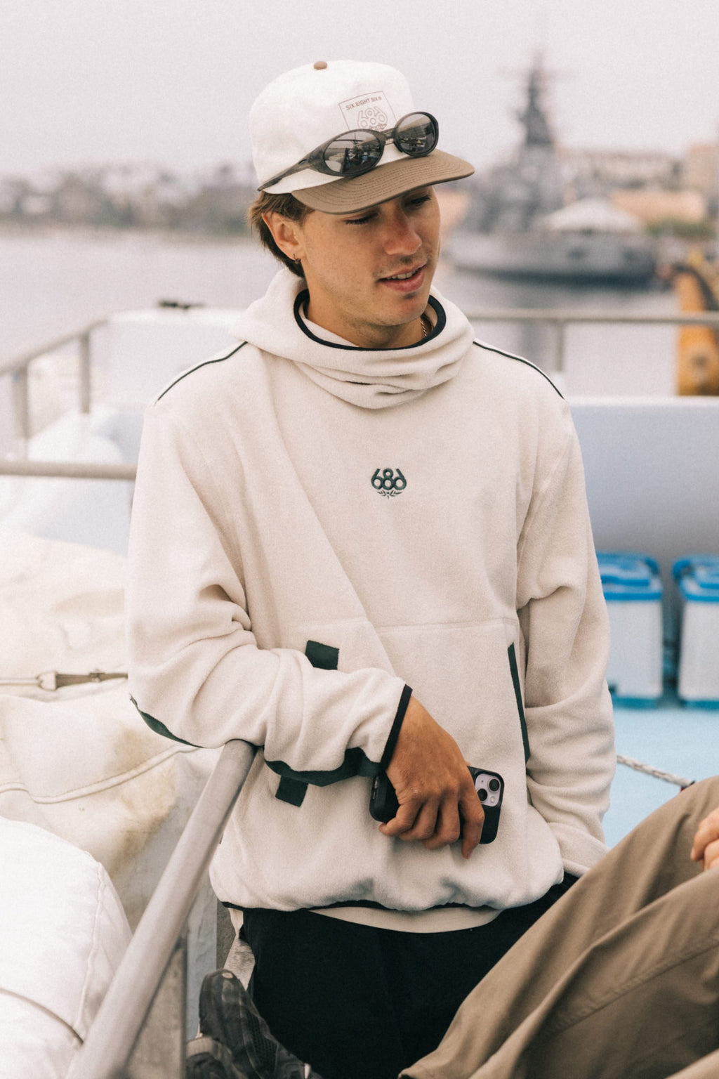 A young man wearing the 686 Mens Clava Fleece Hoody and a cap with sunglasses on top leans against a boat railing, holding a phone. Water, blurred boats, and overcast skies form the background.