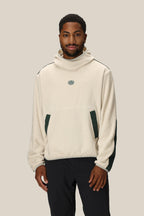 A man wears the 686 Mens Clava Fleece Hoody, a cream polyester sweatshirt by 686 with dark green side accents and a small chest logo, standing against a plain beige background.