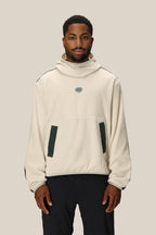 A man wearing the 686 Mens Clava Fleece Hoody, a cream polyester sweatshirt with green details and a small chest logo by 686, stands against a plain beige background.