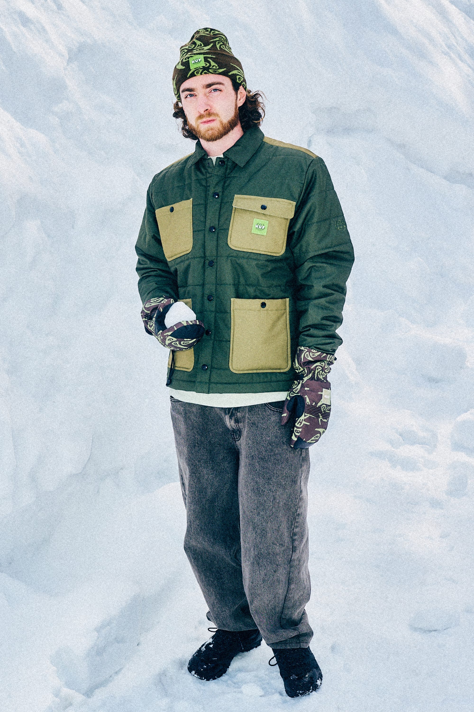 A man stands in the snow wearing a green 686 Mens HUF Puffer Insulated Jacket, a matching beanie, patterned gloves, gray jeans, and black boots. Holding a snowball, he looks at the camera amid snowy scenery.