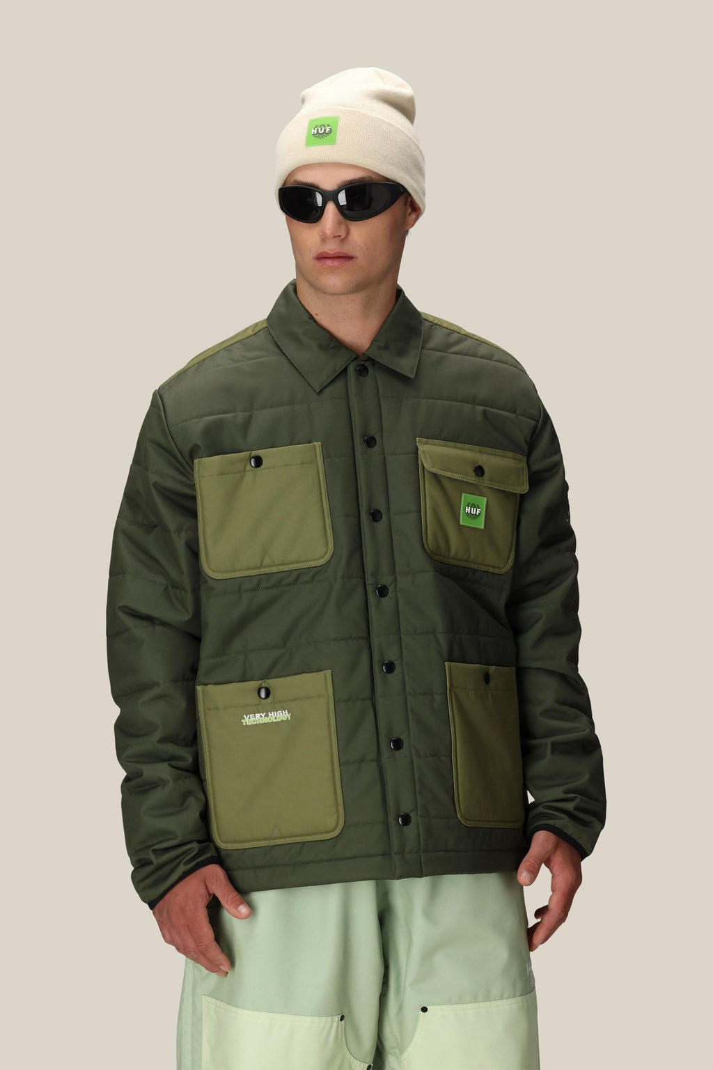 A person wears a cream beanie with a green HUF logo, sunglasses, light green pants, and the 686 Mens HUF Puffer Insulated Jacket by 686, standing against a plain beige background.
