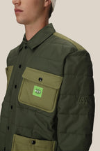 A person wears the 686 Mens HUF Puffer Insulated Jacket in green, featuring large olive pockets, a chest pocket with a small HUF logo patch, and 68 embroidered on the sleeve. The background is plain beige. Brand: 686.