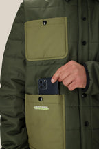 A person in a 686 Mens HUF Puffer Insulated Jacket is placing a black smartphone into a large front pocket. The olive green jacket, by 686, features two big buttoned pockets and a visible VERY HIGH TECHNOLOGY logo.
