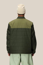 A person is seen from the back wearing a light beige beanie and the 686 Mens HUF Puffer Insulated Jacket by 686, paired with light green pants against a plain beige background.