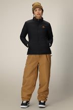 A person wears a black 686 Womens Thermal Puff Insulated Jacket, tan beanie, loose tan pants, and black skate shoes, standing with hands in pockets against a plain beige background.