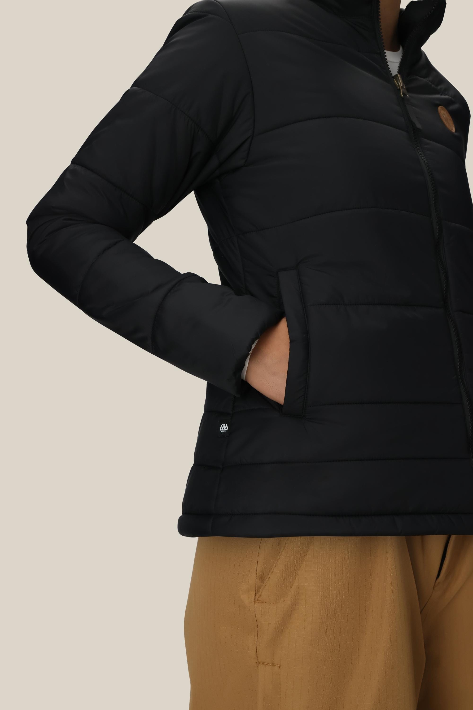 A woman wears the 686 Womens Thermal Puff Insulated Jacket in black polyester with a chest logo and zipper, right hand in pocket, paired with tan pants against a light beige background.